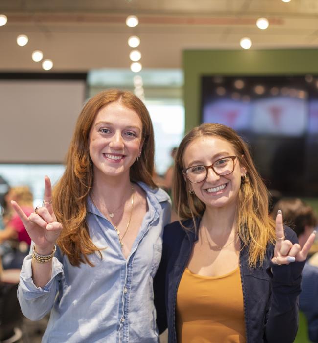 Two students smile and hold up a "hook 'em" hand sign. 
