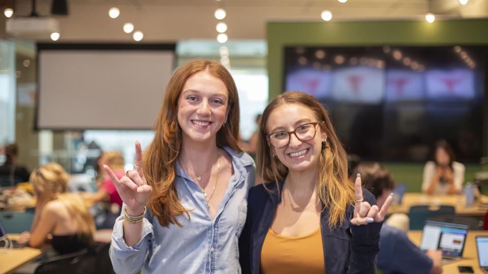 Two students smile and hold up a "hook 'em" hand sign. 